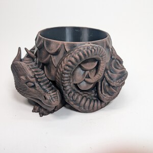 Pode incluir: Um vaso de plantas castanho com tema de drag&atilde;o e interior escuro. O vaso apresenta um design detalhado da cabe&ccedil;a e cauda do drag&atilde;o, com escamas texturizadas e uma cauda enrolada. O drag&atilde;o est&aacute; em posi&ccedil;&atilde;o de repouso, adicionando um elemento de fantasia &agrave; decora&ccedil;&atilde;o.