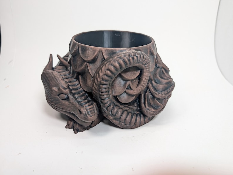 Pode incluir: Um vaso de plantas castanho com tema de drag&atilde;o e interior escuro. O vaso apresenta um design detalhado da cabe&ccedil;a e cauda do drag&atilde;o, com escamas texturizadas e uma cauda enrolada. O drag&atilde;o est&aacute; em posi&ccedil;&atilde;o de repouso, adicionando um elemento de fantasia &agrave; decora&ccedil;&atilde;o.
