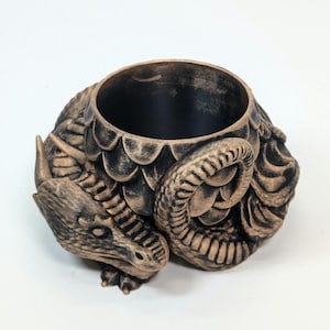 Pode incluir: Um recipiente com tema de drag&atilde;o em bronze com uma abertura circular. O recipiente apresenta um design de drag&atilde;o detalhado, incluindo escamas, uma cabe&ccedil;a e uma cauda enrolada. O interior &eacute; preto e a est&eacute;tica geral &eacute; inspirada na fantasia.