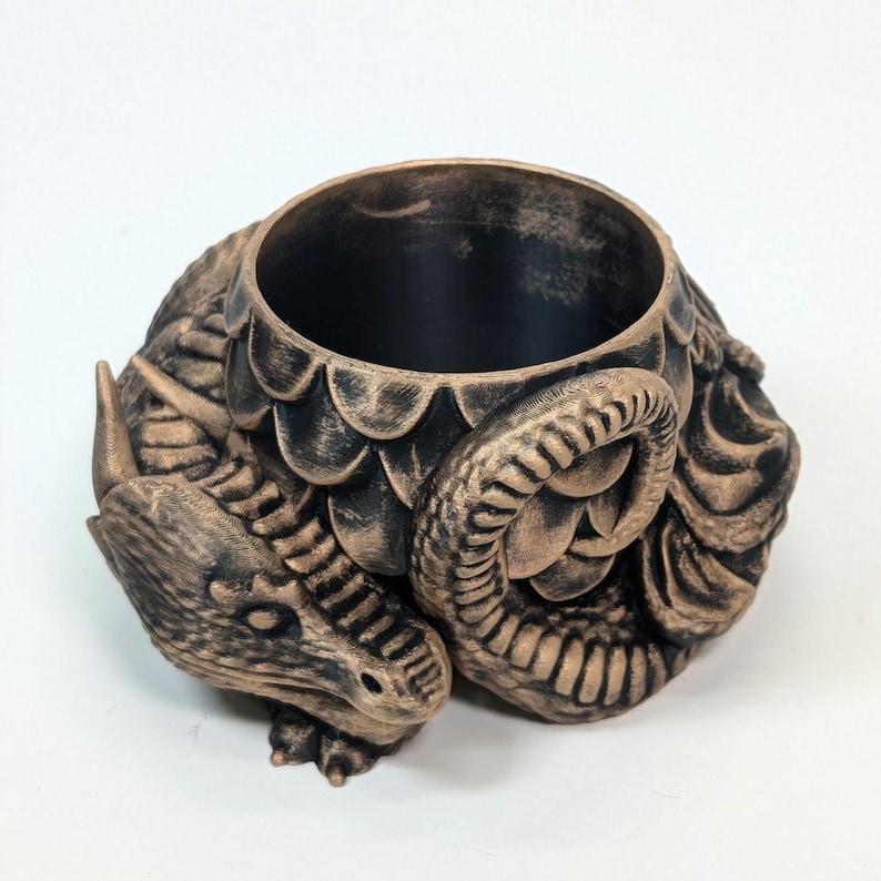Pode incluir: Um recipiente com tema de drag&atilde;o em bronze com uma abertura circular. O recipiente apresenta um design de drag&atilde;o detalhado, incluindo escamas, uma cabe&ccedil;a e uma cauda enrolada. O interior &eacute; preto e a est&eacute;tica geral &eacute; inspirada na fantasia.