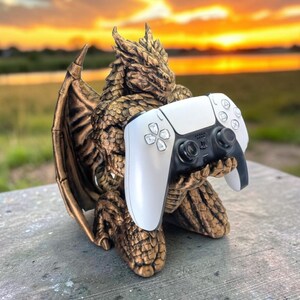 Puede incluir: Una figura de dragón color bronce con escamas texturizadas y alas plegadas, arrodillada mientras sostiene un mando de videojuego inalámbrico blanco y negro en sus manos. El dragón tiene una cabeza y un cuerpo detallados, sirviendo como soporte.