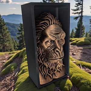 Può includere: Un busto color bronzo di una figura di Bigfoot è esposto all'interno di una cornice rettangolare nera. La scultura presenta dettagliati lineamenti del viso e lunghi capelli. La cornice è ambientata sullo sfondo di una superficie muschiosa e di una vista panoramica sulle montagne.