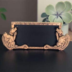 May include: Ornate bronze-colored picture frame with a black insert. The frame features detailed dragon figures on each side and decorative scrollwork along the top and bottom edges. A decorative home decor item.