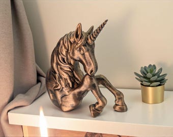 May include: A bronze-colored unicorn figurine in a seated position. The mythical creature has a detailed mane, a spiraled horn, and is posed with its head resting on its front legs. A small succulent plant in a gold pot is next to the unicorn.