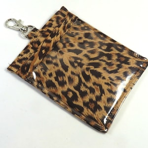 Puede incluir: Un llavero de estampado de leopardo marrón y negro con una ventana de plástico transparente para guardar una tarjeta o un documento de identidad. El llavero tiene un clip de metal plateado.