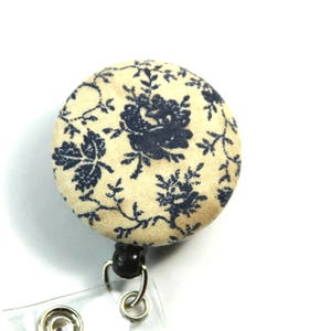 May include: A cream-coloured retractable badge holder with a blue floral pattern. The holder has a clear plastic badge holder and a black swivel clip.