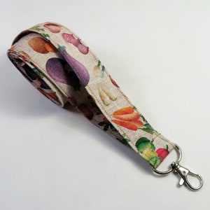 May include: A beige fabric lanyard with a silver key ring. The lanyard is printed with a pattern of colorful vegetables, including carrots, onions, and eggplant.