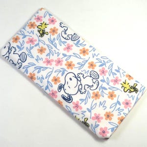 May include: A rectangular, folded fabric item featuring a repeating pattern of Snoopy, Woodstock, pink and orange flowers, and blue leaves on a white background. The design is a whimsical and colorful illustration.