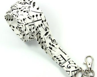 May include: White lanyard with a black musical note pattern. The lanyard has a metal clasp and key ring. The fabric is folded over itself. The lanyard is designed to hold keys or an ID badge.