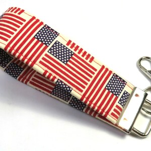 May include: A white fabric key chain with a red, white, and blue American flag pattern. The key chain has a silver metal key ring and a silver metal clasp.