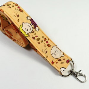 Könnte beinhalten: Ein gelber Lanyard mit einem Peanuts-Charakterdruck mit Charlie Brown und Snoopy. Der Lanyard hat einen silbernen Drehverschluss.