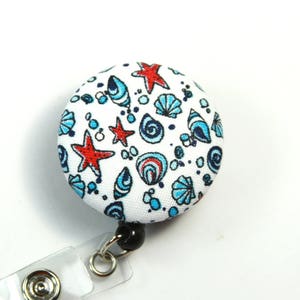 May include: A white button with a blue and red seashell and starfish pattern. The button is attached to a retractable badge holder.