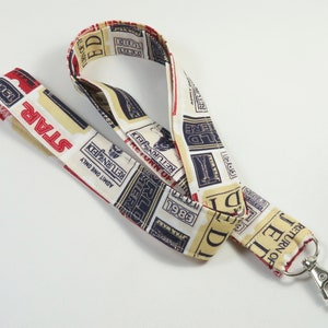 Pode incluir: Um cordão branco, vermelho e azul com tema Star Wars. O cordão tem um clipe prateado e apresenta um padrão repetido de recortes de jornais com o texto "O Retorno de Jedi" e "1983".