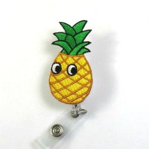 Könnte beinhalten: Eine gelbe Ananas mit grünen Blättern und Kulleraugen ist an einem durchsichtigen, einziehbaren Ausweishalter aus Kunststoff befestigt.