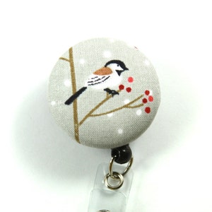 May include: A gray fabric badge reel with a white bird on a branch with red berries and a snowy background.