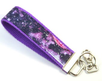May include: A purple wristlet keychain with a galaxy design. The wrist strap is a solid purple color, and the center features a print of a nebula with pink, purple, and white hues. A silver-tone metal clasp is attached.
