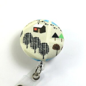 May include: A white fabric button with a black, brown, and green design of a house, trees, and snow. The button is attached to a clear plastic retractable badge holder.