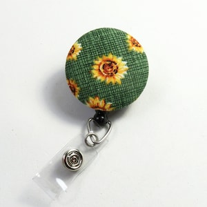 Op de afbeelding: Een groene stoffen knop met een zonnebloemmotief, bevestigd aan een transparante, intrekbare badgehouder van plastic met een zwarte draaibare clip.