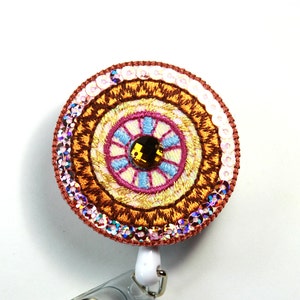 May include: A round, colorful badge reel with a brown, yellow, pink, and blue embroidered design. The center of the design features a large, clear rhinestone.