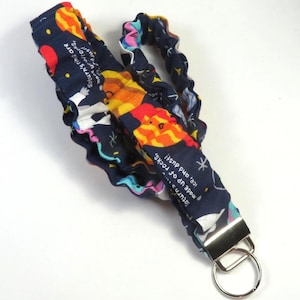 May include: A navy blue lanyard with a repeating pattern of planets and stars. The lanyard has a metal clasp and a key ring. Text on the lanyard reads "Saturn's rings are made up of rocks, ice, and dust!"
