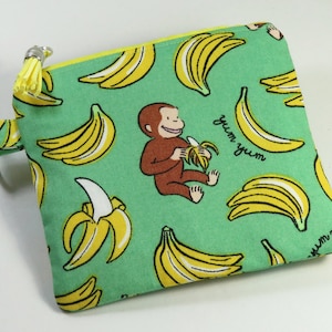 May include: A square, green fabric pouch with a yellow zipper and tassel. The pouch features a repeating pattern of yellow bananas and a brown monkey eating a banana. The words "yum yum" are also printed on the pouch.