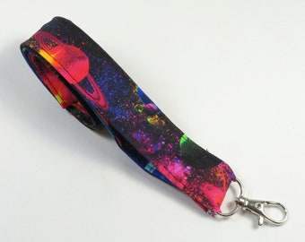 May include: A black fabric lanyard with a colorful space-themed print featuring planets, stars, and nebulae. The lanyard has a silver metal clip.