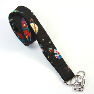 May include: Black lanyard with a space-themed print featuring rockets, planets, and stars. The lanyard has a silver swivel clip.