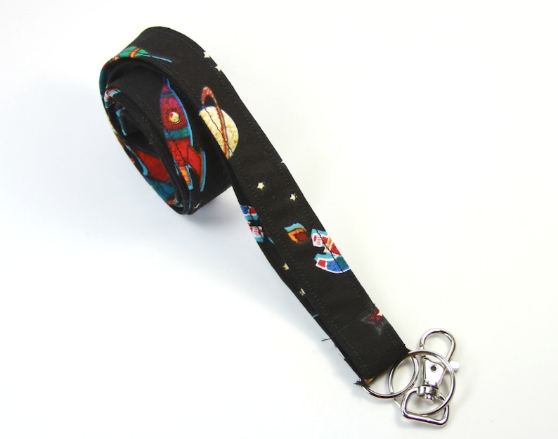 May include: Black lanyard with a space-themed print featuring rockets, planets, and stars. The lanyard has a silver swivel clip.