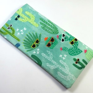 May include: A green fabric eye mask with a pattern of cacti wearing sunglasses. The cacti are green, yellow, and white. The background is a light green watercolor wash.