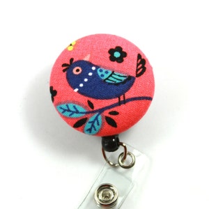 May include: A pink fabric button with a blue bird design, a green branch, and black flowers. The button is attached to a clear plastic retractable badge holder with a black clip.