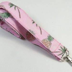 Peut inclure: Lanière rose avec un motif d'ananas et de palmiers. La lanière est munie d'un mousqueton pivotant argenté.