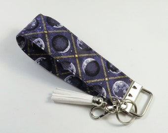 May include: A purple and gold fabric key chain with a white tassel and a silver key ring. The fabric features a repeating pattern of moons and stars.