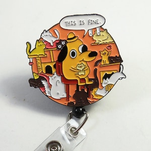 May include: A round badge featuring a yellow dog sitting in a chair, surrounded by cats, with the text "THIS IS FINE" in a speech bubble. The badge has a black retractable reel and a clear plastic strap.