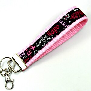 May include: Pink key chain with a black and white ribbon design. The ribbon has the words "Hope", "Love", "Happy Joy" and "Support" written on it.