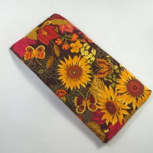Puede incluir: Artículo de tela rectangular con fondo marrón, con un vibrante estampado floral. El diseño incluye grandes girasoles en tonos amarillos y naranjas, junto con mariposas y hojas de otoño en rojo y dorado.