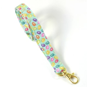 Könnte beinhalten: Ein Stoff-Lanyard mit einem sich wiederholenden Muster aus bunten, oval geformten Designs auf hellblauem Hintergrund. Das Lanyard hat einen goldfarbenen Metallverschluss. Das Muster umfasst orange, gelbe, rosa und lila Formen.