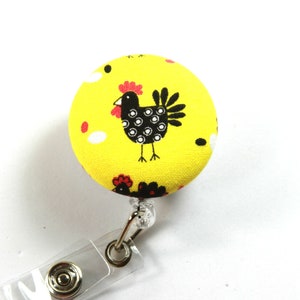 May include: A yellow fabric button with a black and white chicken design and red accents. The button is attached to a clear plastic retractable badge holder.