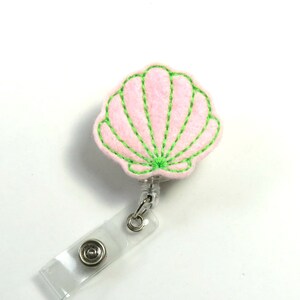 May include: A pink seashell with a green outline, attached to a clear plastic retractable badge holder.
