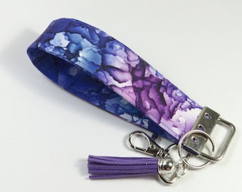 May include: A wristlet key fob with a vibrant watercolor design in shades of blue and purple. It features a silver-tone metal clasp, key ring, and a matching purple tassel. The fabric wrist strap is approximately 1 inch wide.