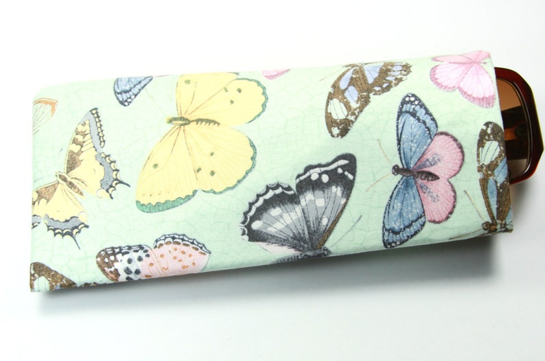 May include: A green fabric eyeglasses case with a butterfly print. The case is closed and has a brown pair of glasses inside.