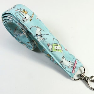 Może przedstawiać: Jasno niebieski sznurek z białym paskiem z tkaniny, na którym powtarza się wzór myszy w kapeluszach trzymających przedmioty. Sznurek ma srebrny karabińczyk obrotowy.