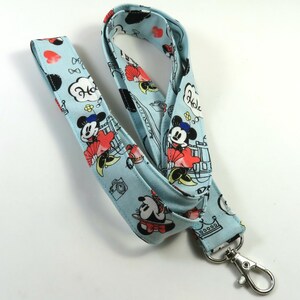 May include: A light blue lanyard with a Mickey Mouse and Minnie Mouse print. The lanyard has a silver swivel clasp. The print features Mickey and Minnie in various poses, including holding hands and kissing. The lanyard also features the words "Hola" and "Hello" in a speech bubble.