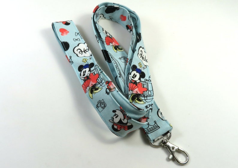 May include: A light blue lanyard with a Mickey Mouse and Minnie Mouse print. The lanyard has a silver swivel clasp. The print features Mickey and Minnie in various poses, including holding hands and kissing. The lanyard also features the words "Hola" and "Hello" in a speech bubble.