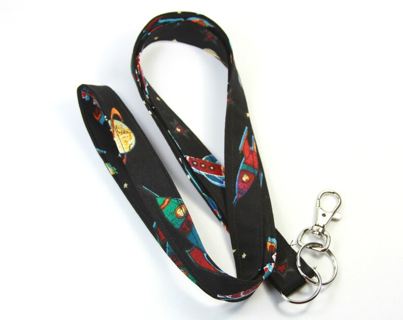 May include: A black lanyard with a space-themed print featuring rockets and planets. The lanyard has a silver swivel clip and a silver key ring.