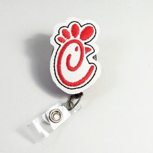 Puede incluir: Logotipo de pollo bordado en blanco y rojo sobre fondo negro, unido a un portagafete de plástico transparente con un clip plateado.