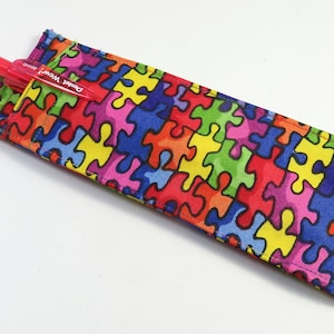 Op de afbeelding: Een kleurrijk stoffen potlood etui met een puzzelstukjes patroon. Het etui heeft een metalen clip en een rode pen erin.