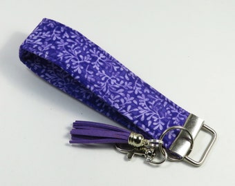 May include: Purple fabric key chain with a white floral pattern and a purple tassel. The key chain has a silver metal ring and a silver metal clasp.