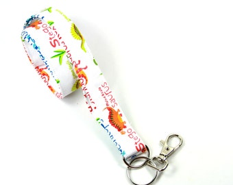 May include: White lanyard with a colorful dinosaur print and the text "Stego" and "saurus" printed on it. The lanyard has a silver metal clip and a silver key ring.