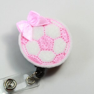 May include: A white soccer ball with pink stitching and a pink bow, attached to a clear retractable badge holder.