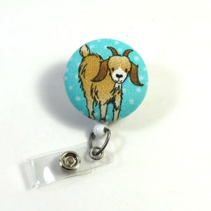 Könnte beinhalten: Ein türkisfarbener Stoffknopf mit weißen Punkten, der eine braune und weiße Comic-Ziege mit braunen Hörnern zeigt. Der Knopf ist an einem durchsichtigen Plastik-Badgehalter mit einem silbernen Metallclip befestigt.